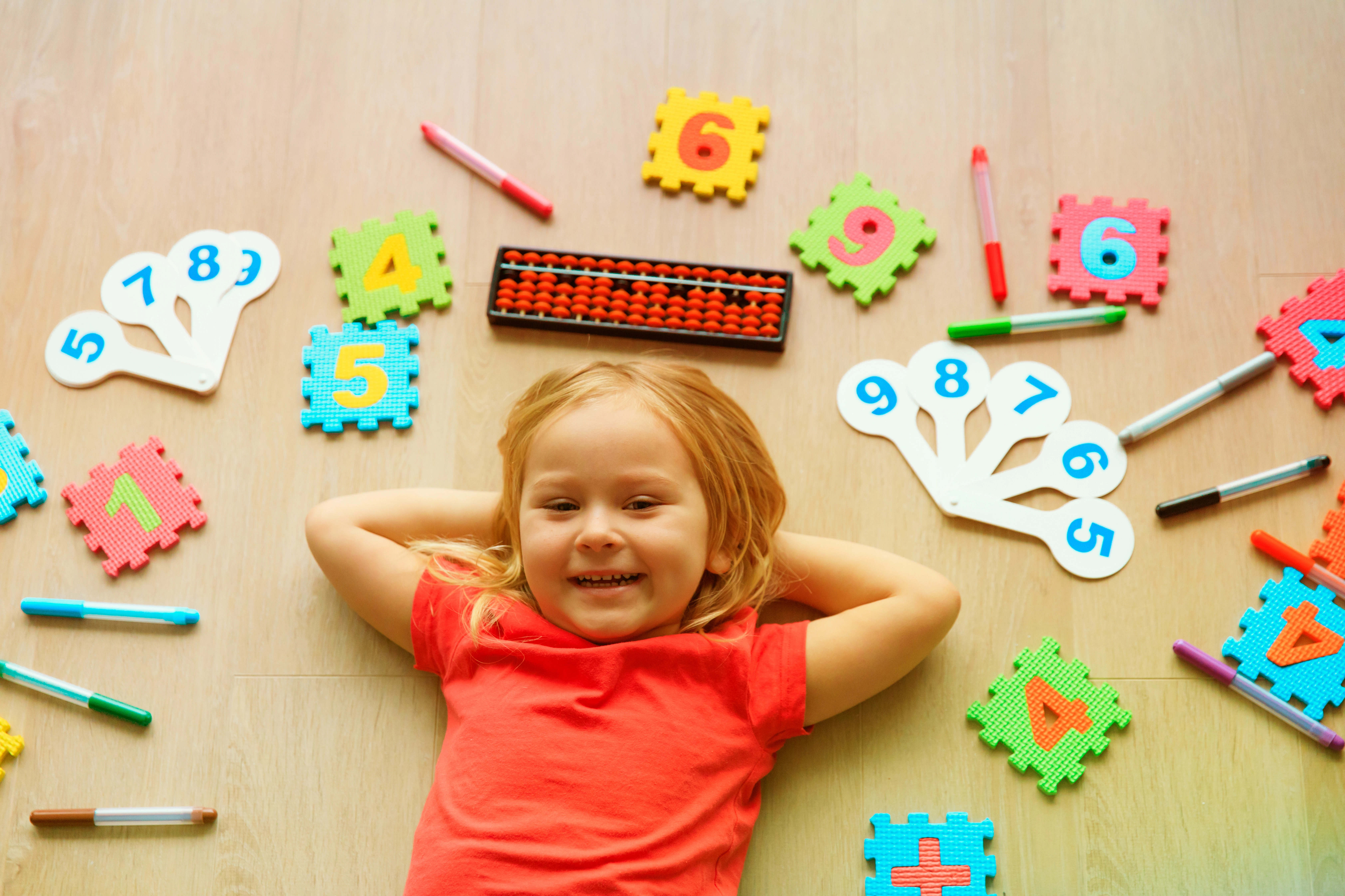A smiling young child in a red shirt lies on the floor surrounded by colorful educational toys, incl