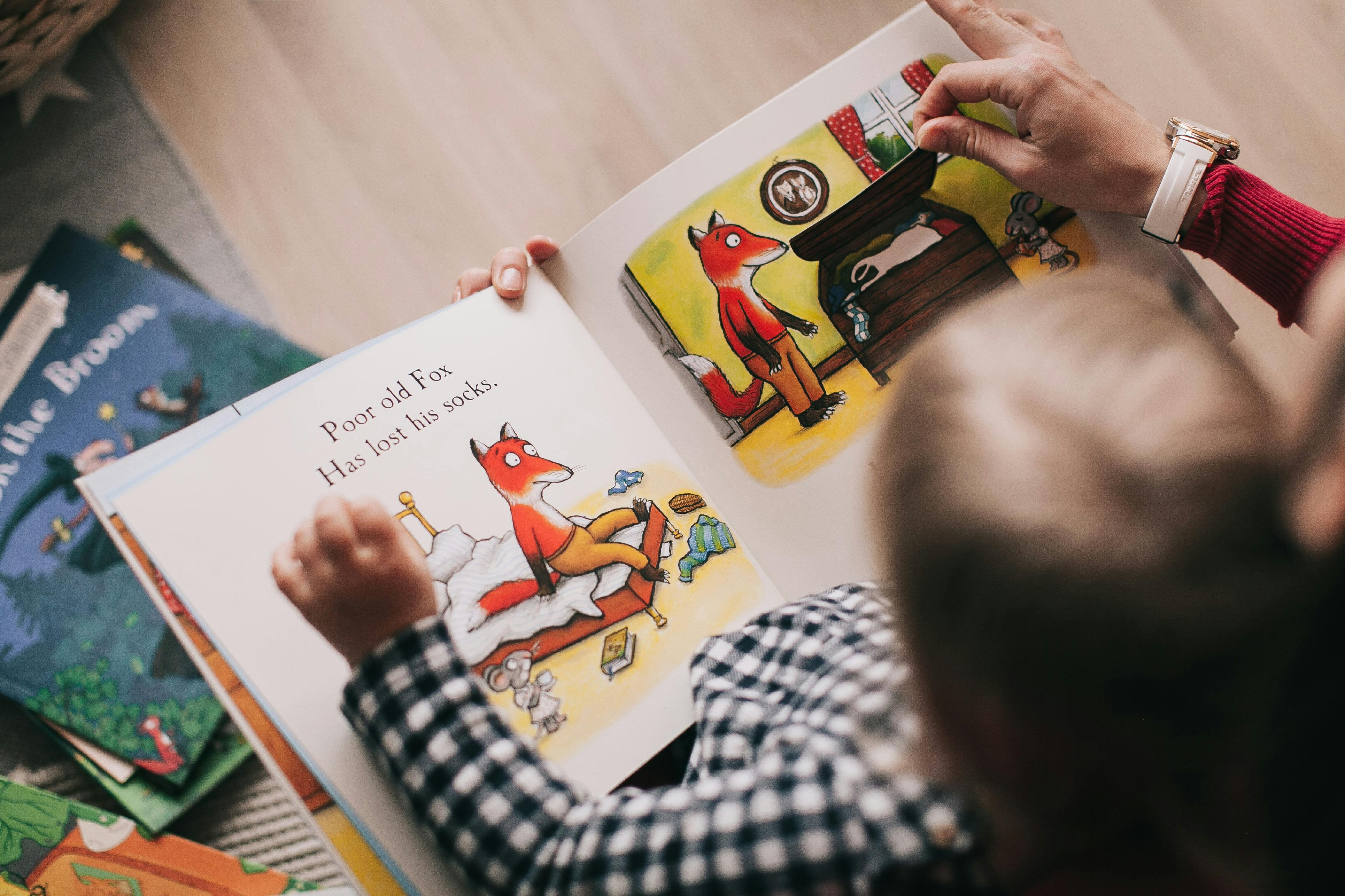 A child in a black-and-white checkered shirt studies English vocabulary with an adult while reading a colorful children’s picture book about a fox who has lost his socks and with only the adult’s supportive hands visible holding the book. 