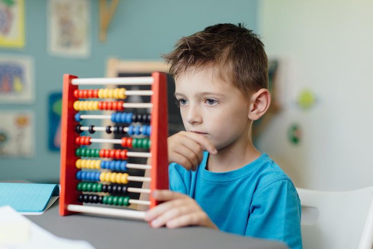 A young boy in a blue t-shirt with a thoughtful expression is using a colorful wooden abacus. He is holding his hand to his chin as if concentrating on a math problem.