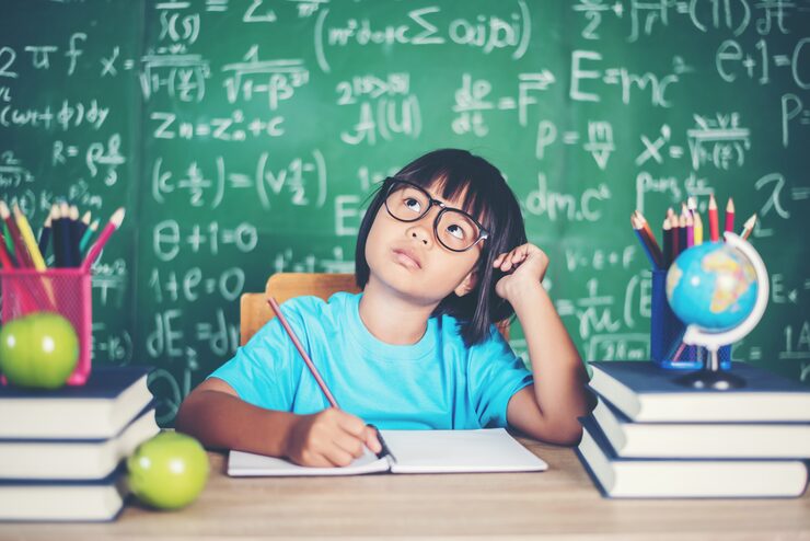 Child thinking about math while writing in a notebook, with chalkboard full of mathematical formulas in the background.