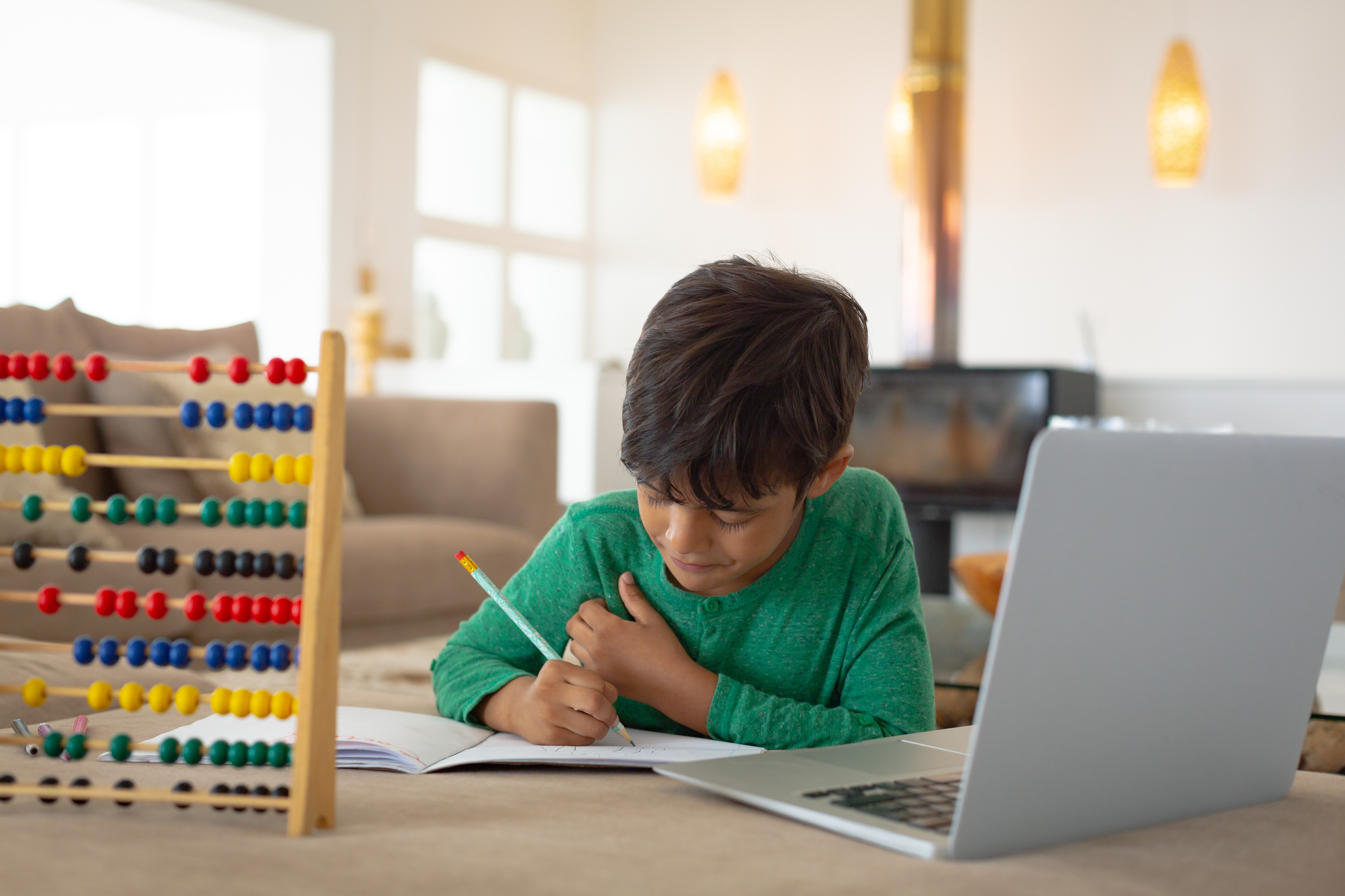 A young boy in a green shirt studying at home using a laptop, writing in a notebook with a pencil and with a colorful abacus for math learning on the table.