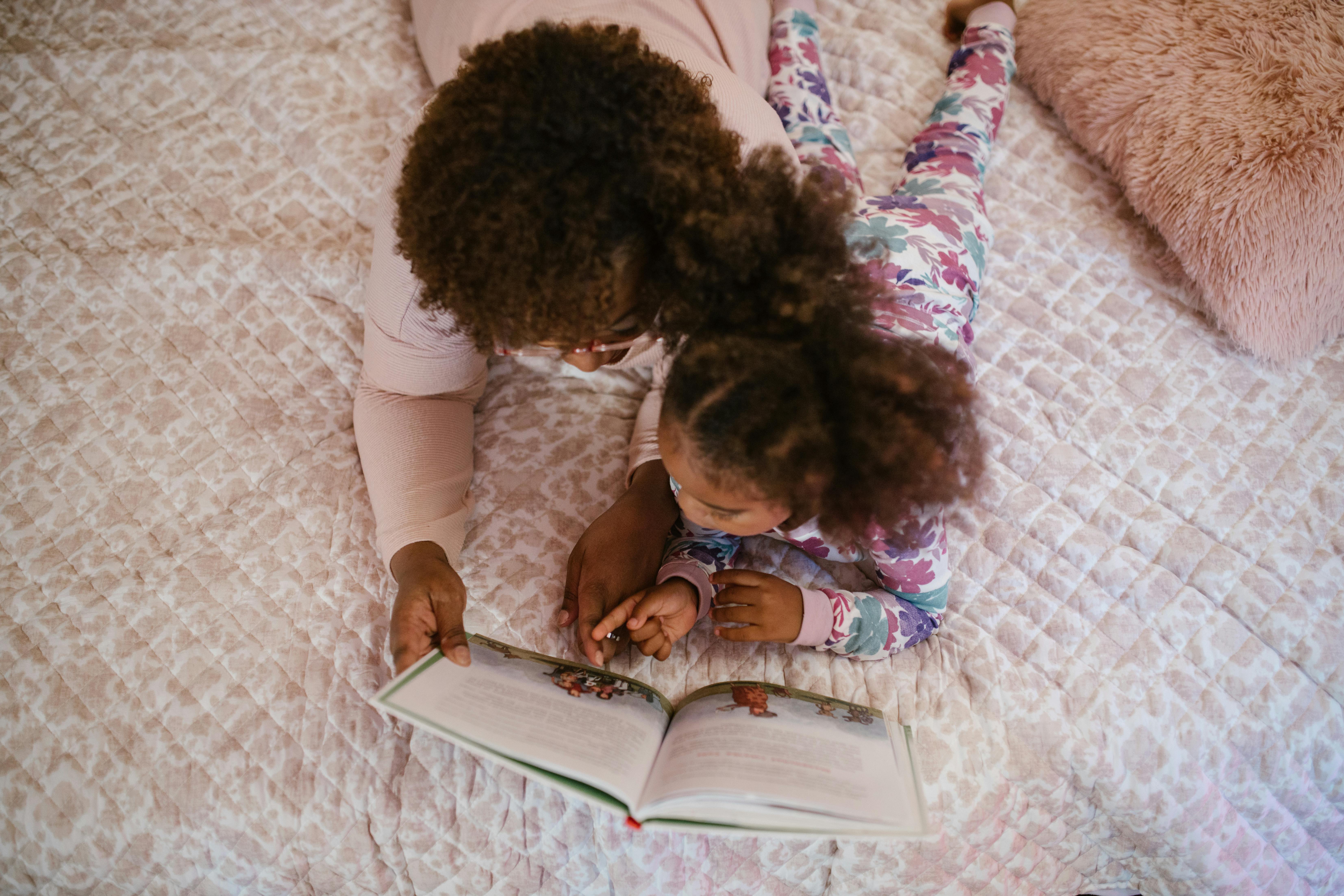 A child in colorful floral pajamas lies on a bed with an adult in a light pink outfit reading a children’s book together. The adult gently points at the words while supporting the child. The child is learning English vocabulary, and the adult helps by reading aloud and guiding understanding.