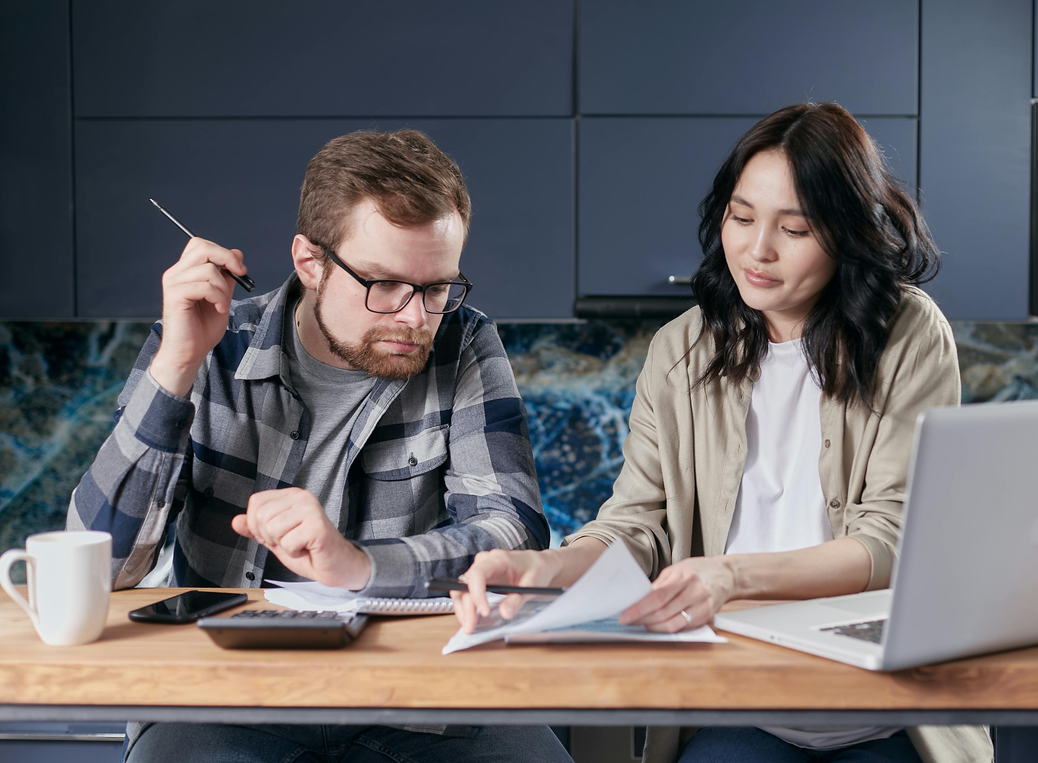 A man and a woman are sitting at a wooden table reviewing their finances or budget. The man on the left has a reddish beard and is wearing a blue and gray flannel shirt over a gray t-shirt. He is holding a pen and has glasses on. The woman on the right has dark hair and is wearing a tan or light brown jacket over a white t-shirt. There are a calculator, laptop, and paperwork on the table, which are tools for their calculation and financial planning.