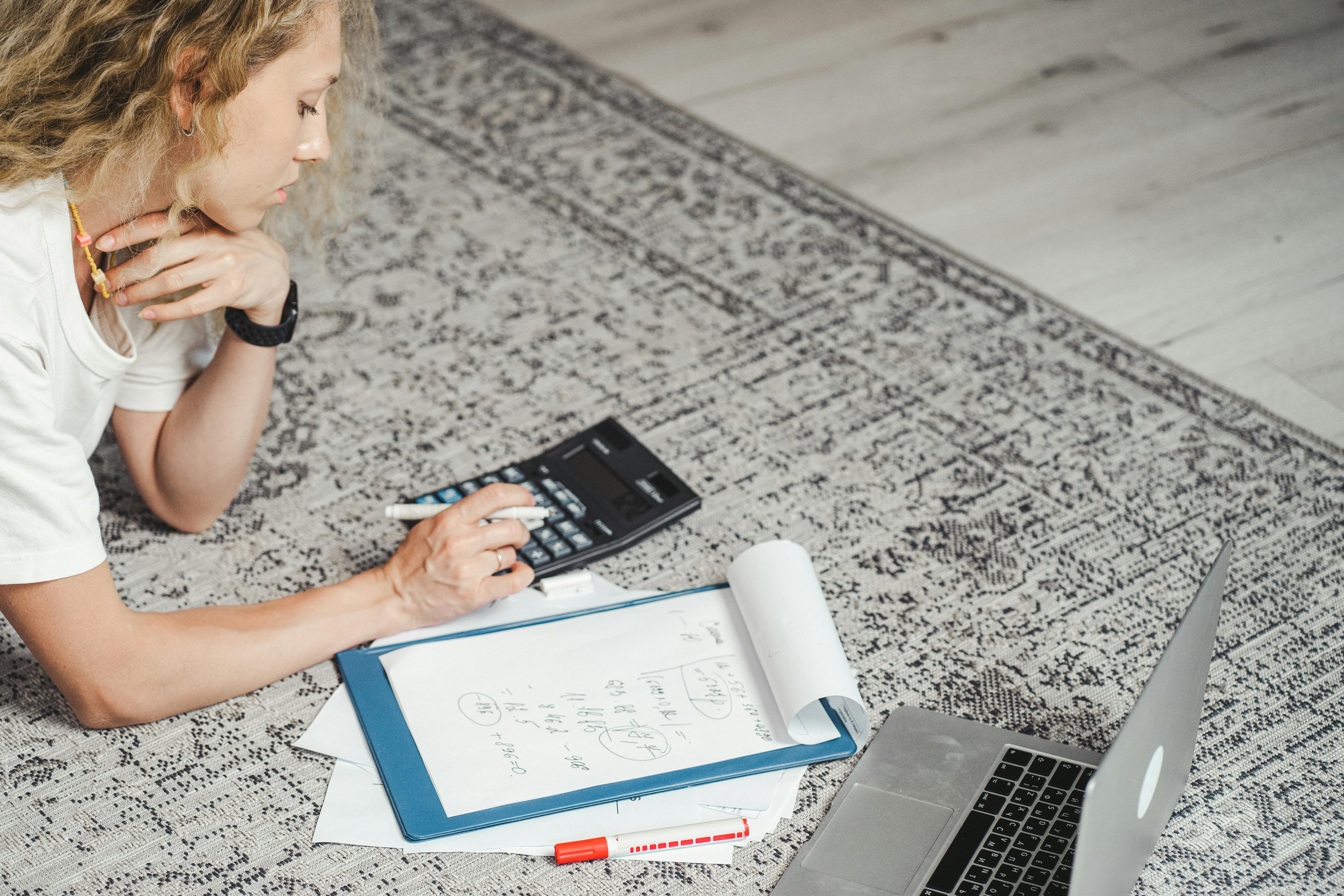 A woman in a white t-shirt is lying face down on a patterned rug engrossed in a math calculation. She is using a calculator and a pen, with a clipboard full of equations and a laptop laid out on the floor in front of her. Her concentration suggests she is working hard.