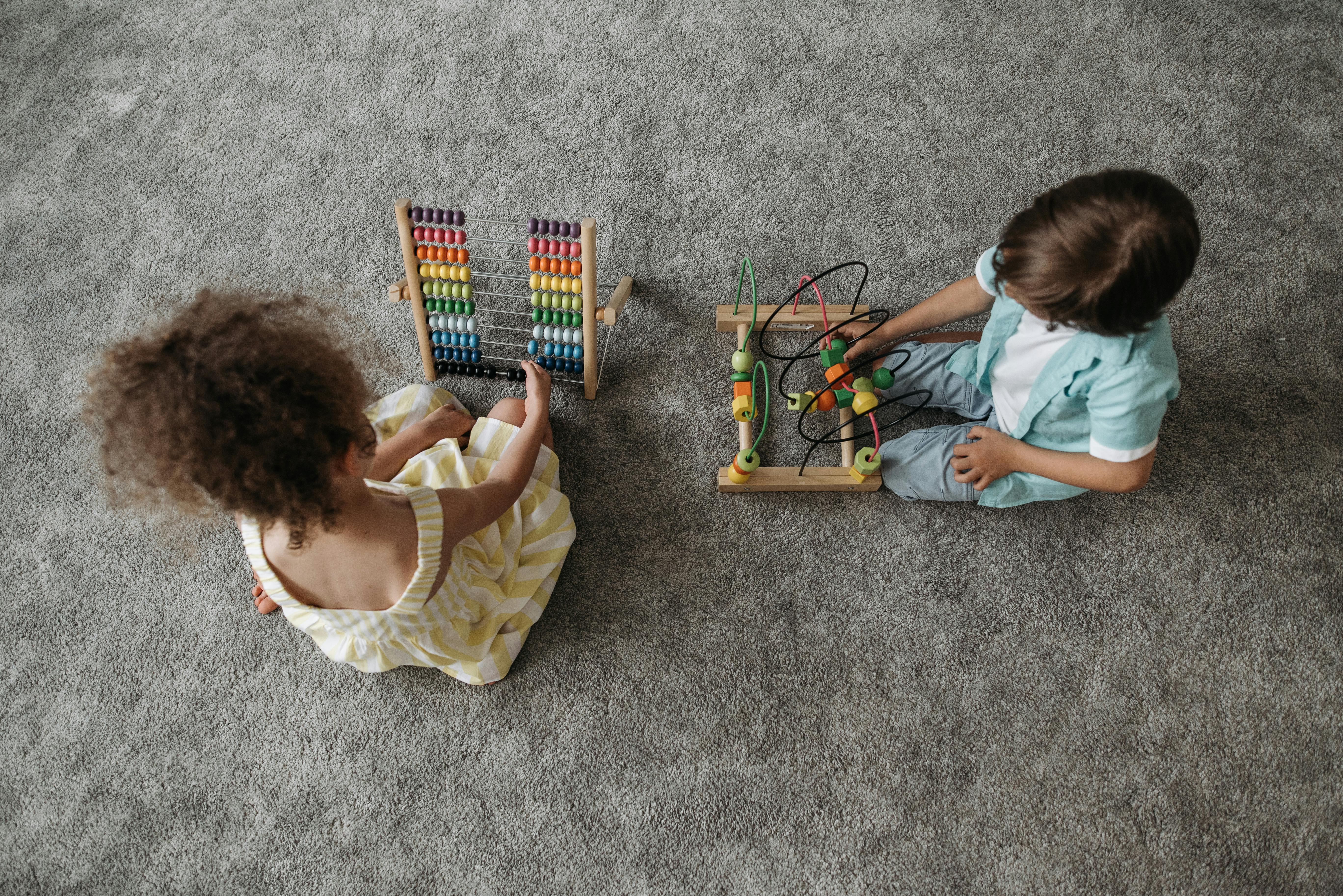 Overhead shot of two young children sitting on a gray carpet, engaged in educational play. One child, wearing a yellow dress is interacting with a large wooden rainbow abacus for counting and math learning. The other child, wearing a blue shirt, is playing with a wooden bead maze or wire puzzle that develops fine motor skills and coordination. The image highlights early childhood development and Abacus training.