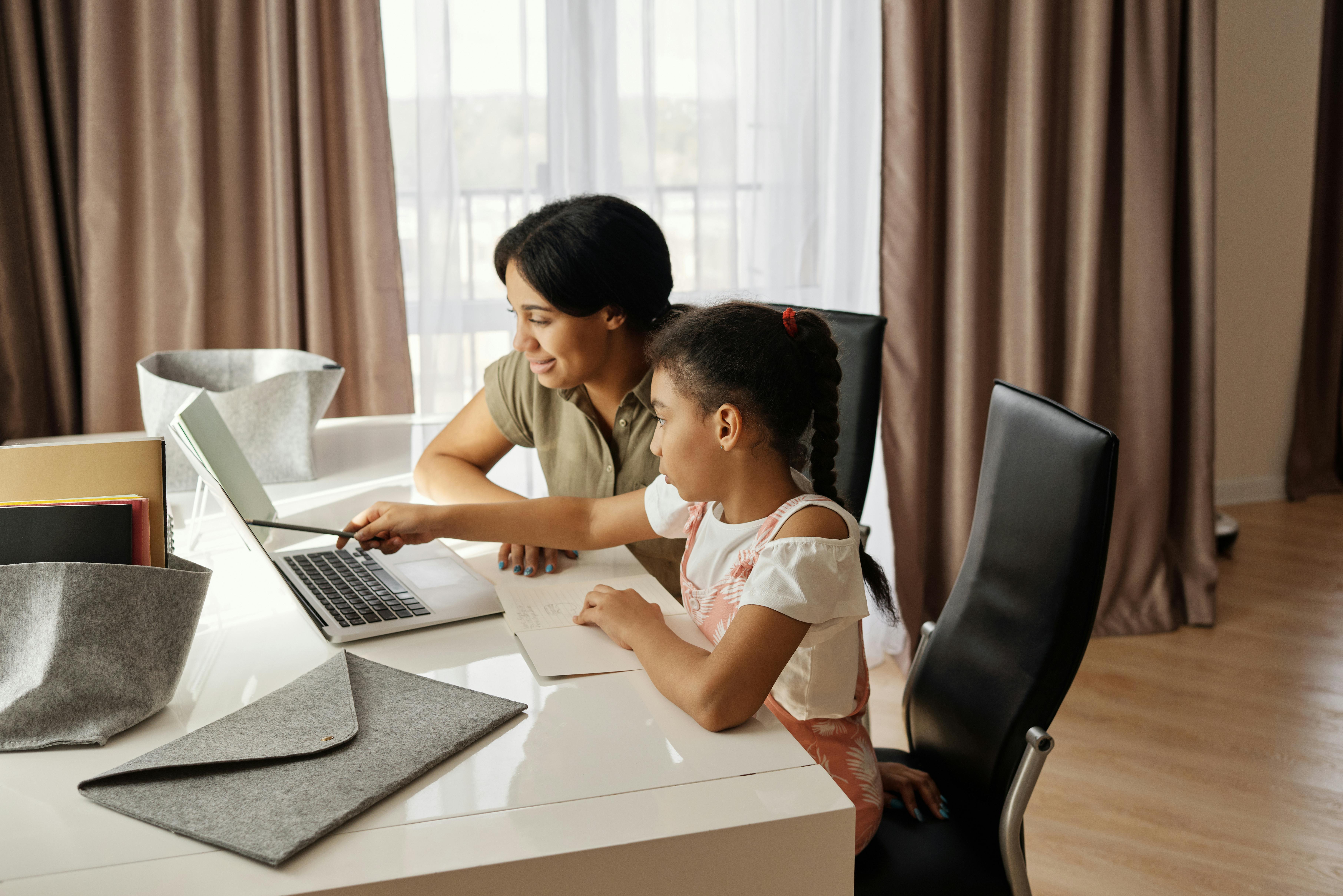 A smiling mother and her young daughter are seated at a white desk, engaged in learning on a laptop. The woman points at the screen with a pen while the girl looks on and takes notes in a notebook. This scene represents modern mental math skills practice and a brain development program, potentially related to Abacus training or Abacus learning at home or in an educational setting.
