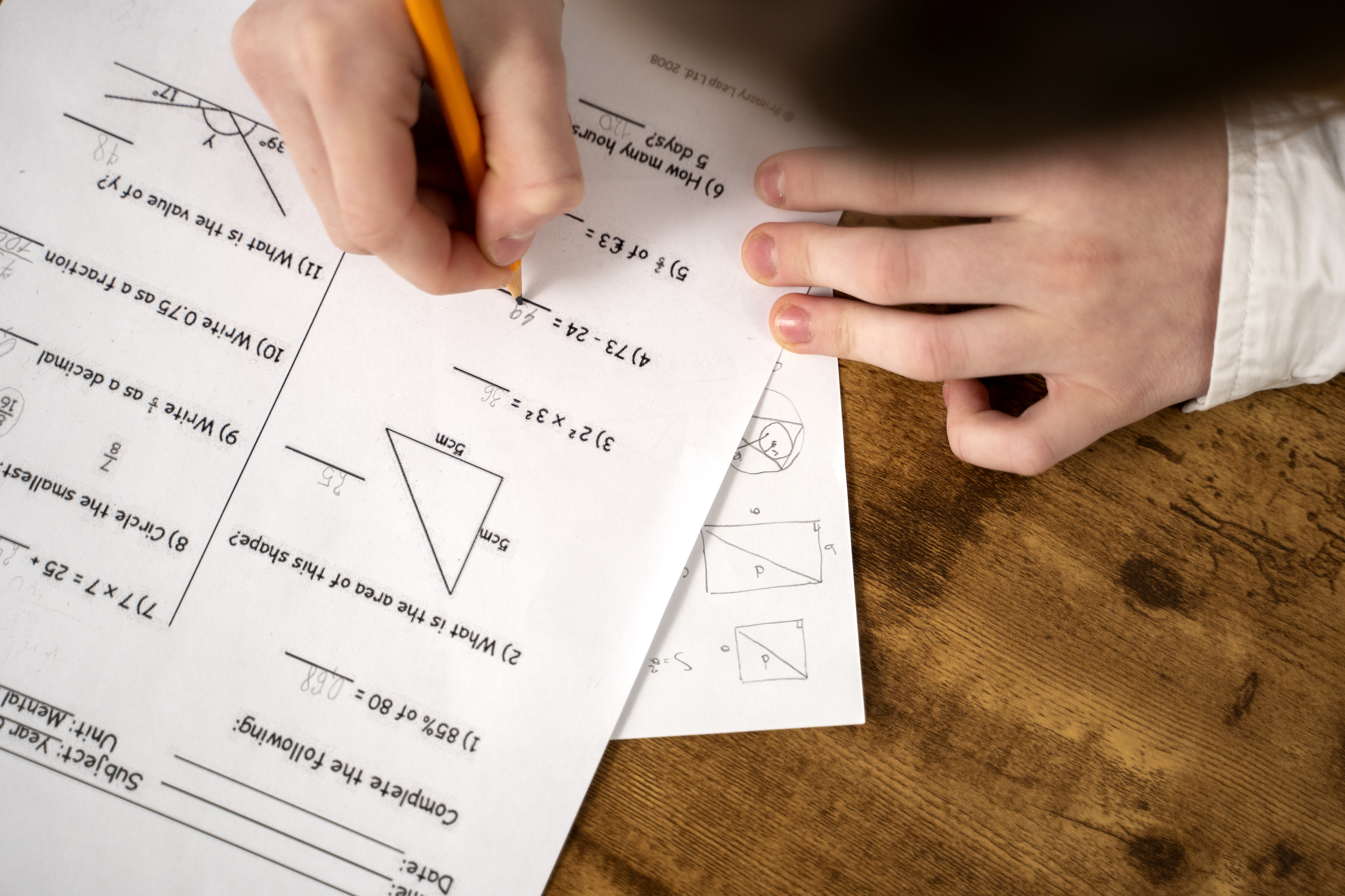Child's handwriting answers on a school math test with problems involving arithmetic and geometry, while their other hand obscures a hidden paper underneath, possibly containing Vedic Math shortcuts or quick calculation notes.